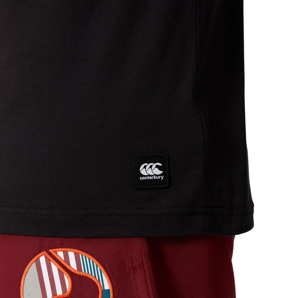 Close-up of the Canterbury Uglies Logo T-shirt—a black cotton-blend tee with the Canterbury logo tag—paired with red shorts featuring a vibrant abstract pattern.