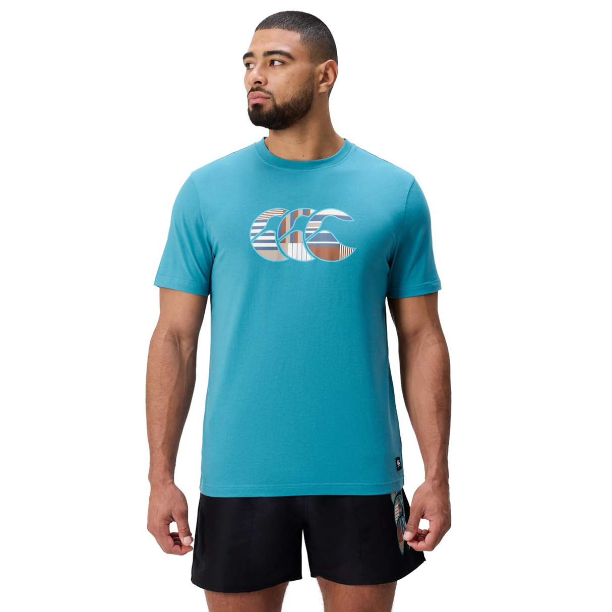 A man stands facing forward with arms at his sides, wearing a teal Canterbury Uglies Logo T-shirt in a soft cotton blend featuring a large logo on the chest, paired with black athletic shorts.
