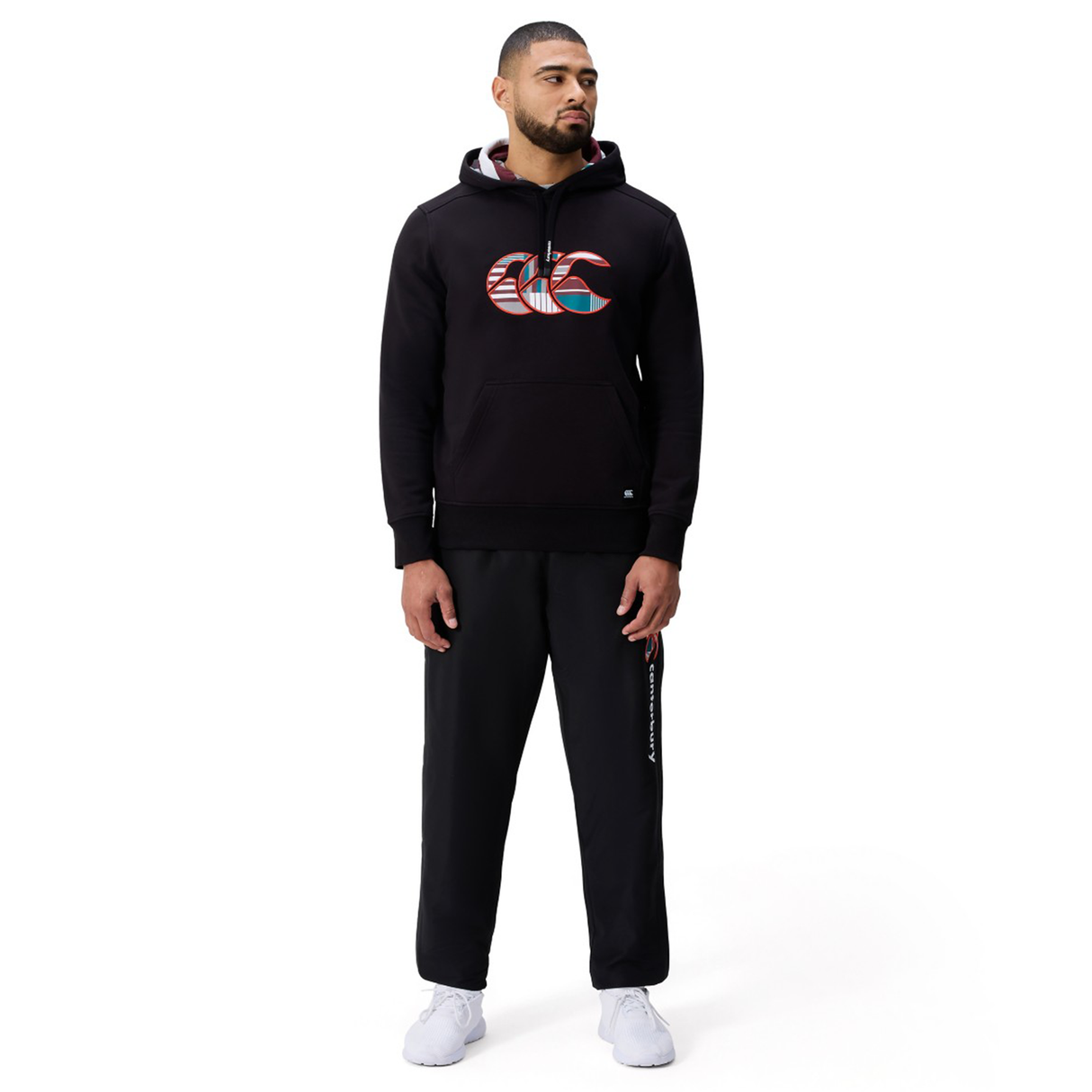 A man stands against a plain white background wearing the Canterbury Uglies Logo Hoodie in black by Canterbury, paired with black sweatpants and white sneakers.