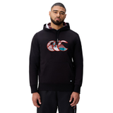 A man wearing a Canterbury Uglies Logo Hoodie in black with a colorful chest graphic and black pants stands against a white background.