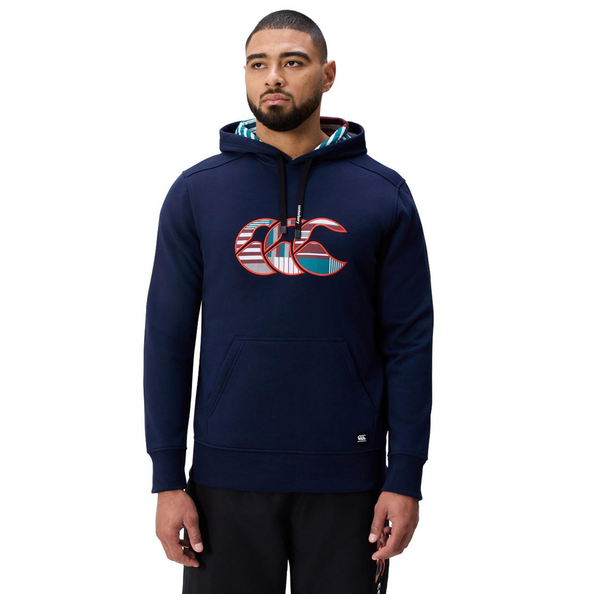 A man wearing a Canterbury Uglies Logo Hoodie in navy blue, featuring the patterned Canterbury logo on the chest, paired with black pants and standing against a white background.
