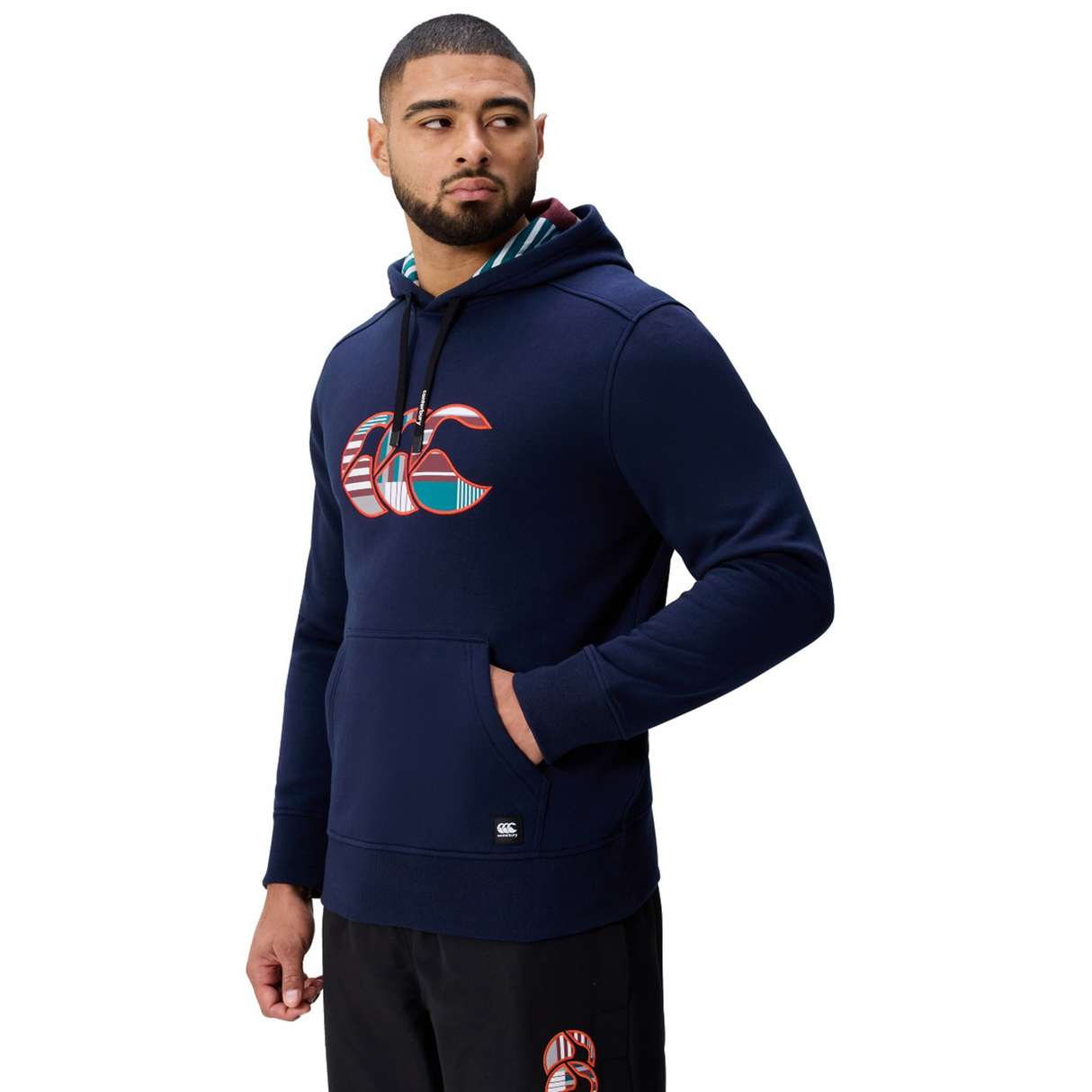 A man in a navy blue Canterbury Uglies Logo Hoodie and black pants stands against a white background, looking to the side.