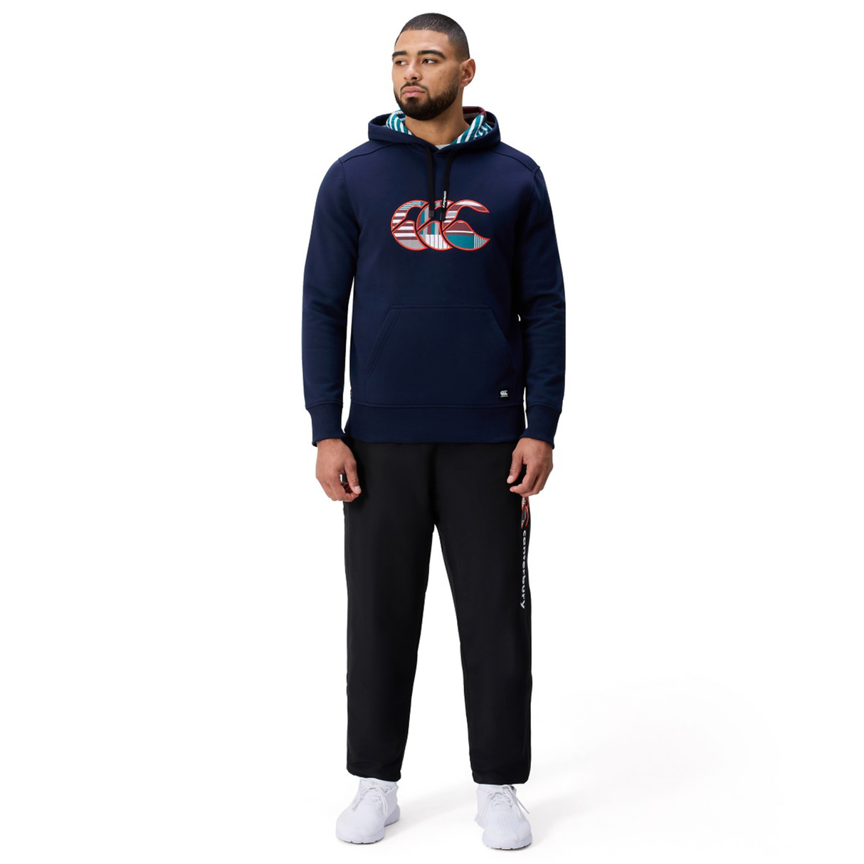 A man wears the Canterbury Uglies Logo Hoodie by Canterbury in navy blue, paired with black pants and white sneakers, standing against a plain white background.