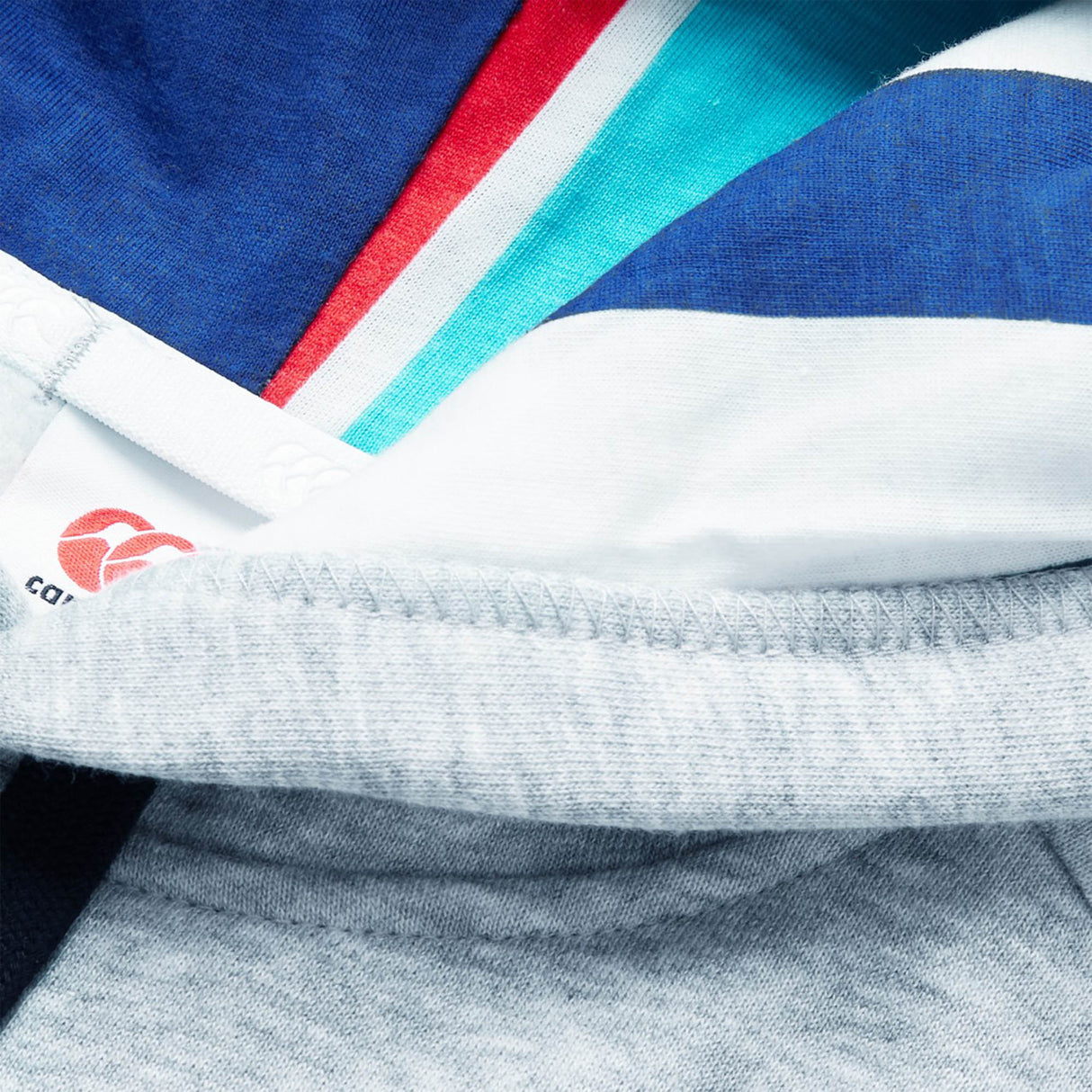 A close-up view of colorful layered fabric—featuring blue, teal, red, white, and gray segments reminiscent of the vibrant designs found on a Canterbury Uglies Logo Hoodie by Canterbury—with a partially visible clothing tag displaying printed text and the distinctive red Uglies Logo.