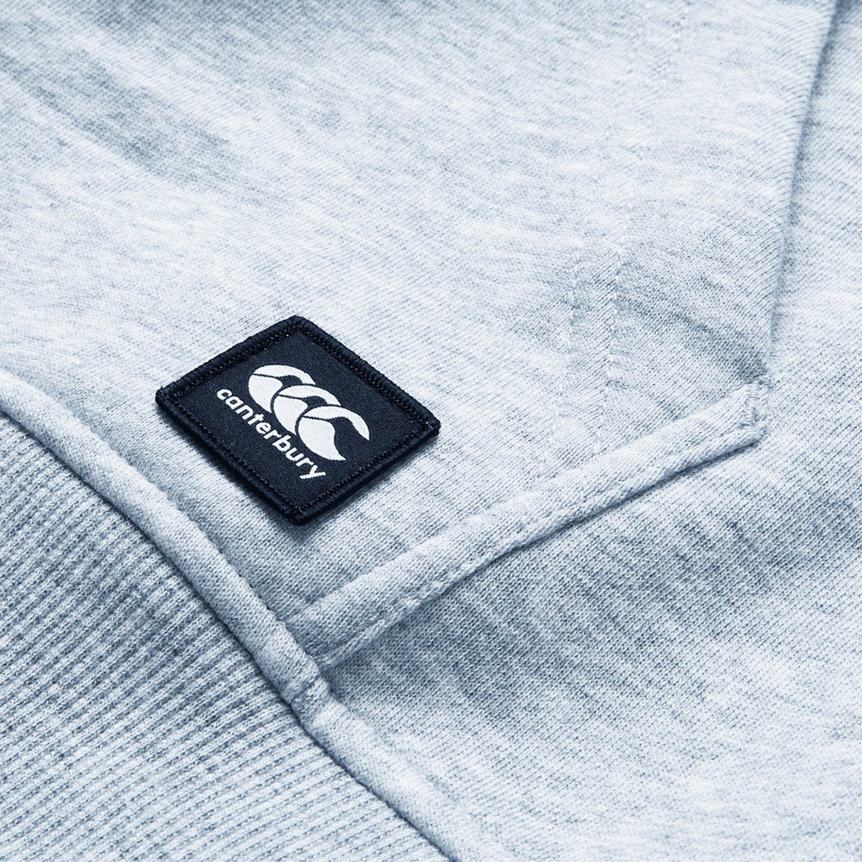 Close-up of a light gray Canterbury Uglies Logo Hoodie displaying a stitched Canterbury logo patch with three white overlapping C's on a black square background.