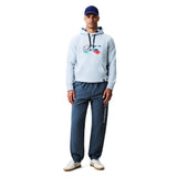 A man stands against a plain white background wearing a blue baseball cap, the Canterbury Uglies Logo Hoodie in gray with a colorful logo design by Canterbury, blue track pants, and white sneakers.