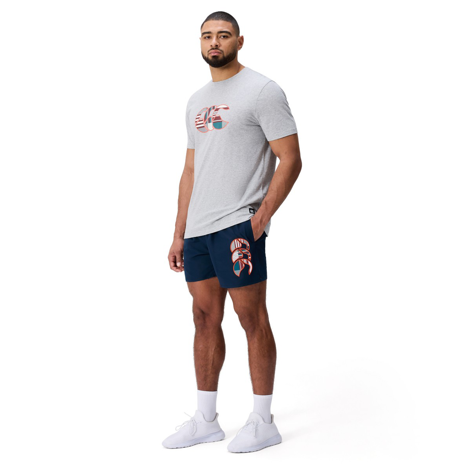 A man faces forward in a light gray T-shirt, Canterbury Uglies 5" Tactic Short by Canterbury with a graphic, white socks, and white sneakers against a plain white background.