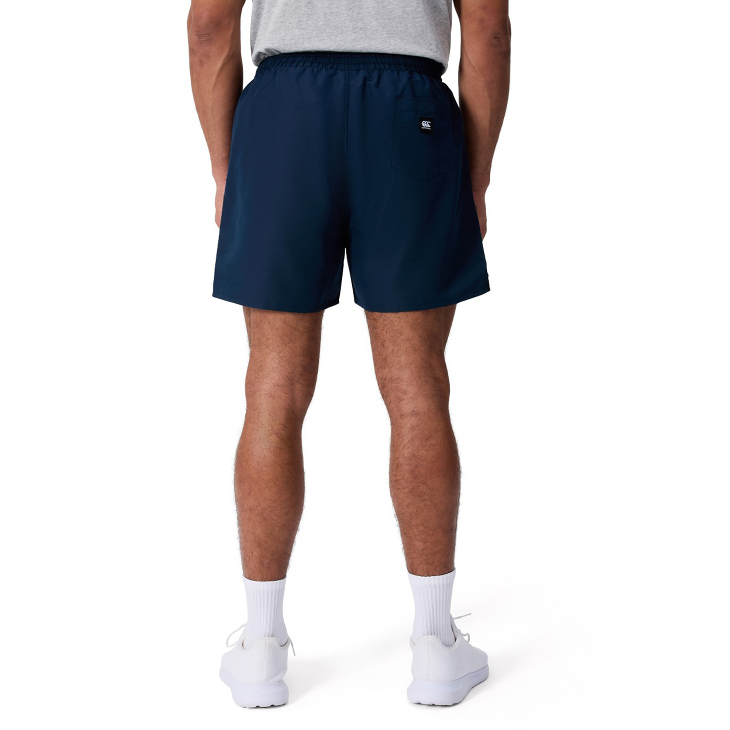 A person is shown from the back against a plain white background, wearing a gray t-shirt, Canterbury Uglies 5" Tactic Shorts in navy blue, white calf socks, and white shoes.