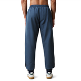 A man is seen from the back wearing Canterbury's Uglies 32" Stadium Pant in blue, featuring an elastic waistband and cuffs, paired with white sneakers and no shirt—perfect for a casual day or post-rugby relaxation.