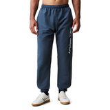 A person wearing blue Uglies 32" Stadium Pant by Canterbury with a logo on the left thigh and white sneakers stands against a white background.