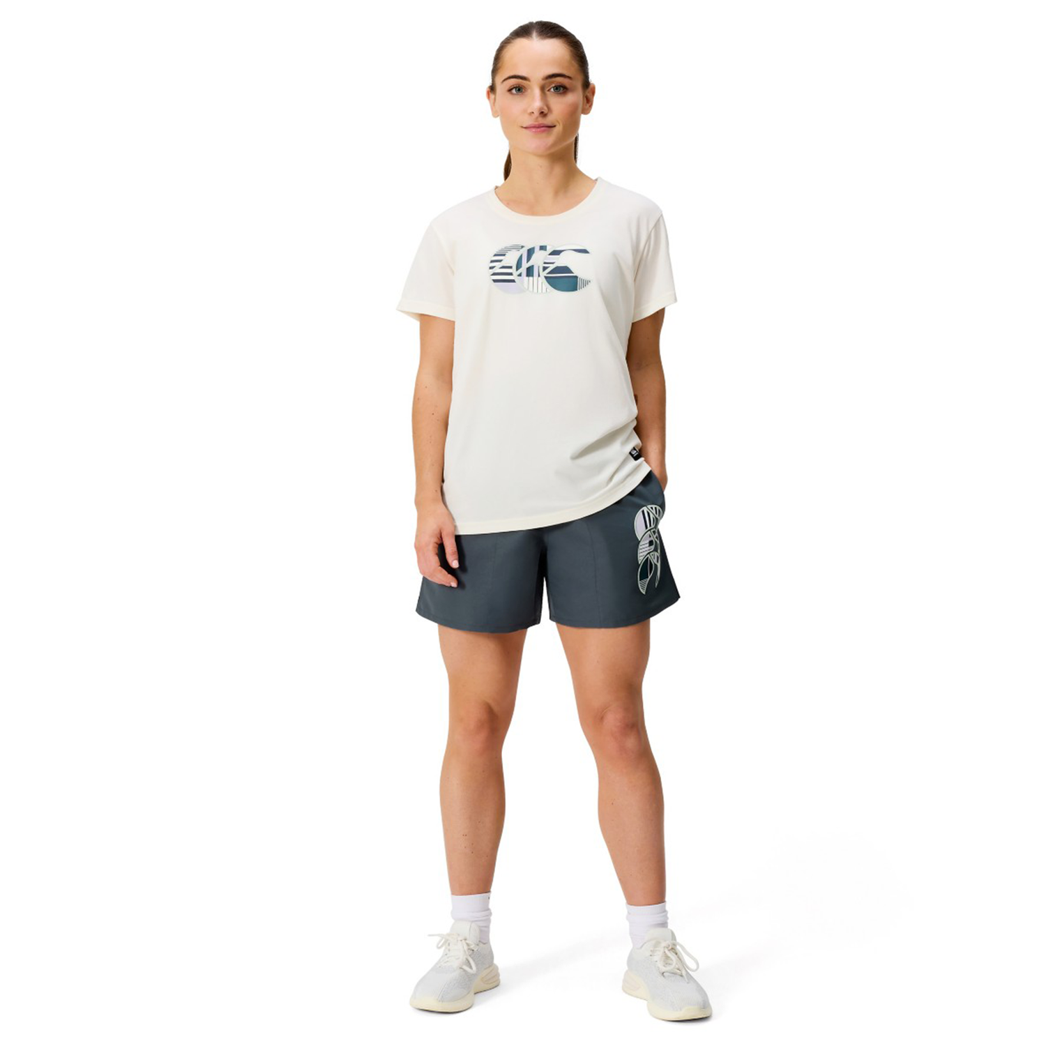 A woman faces forward against a white background wearing a white graphic t-shirt, Canterbury Women’s Canterbury Uglies Tactic Short in dark color, white socks, and white sneakers.