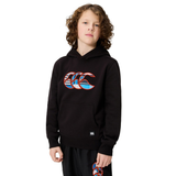 A child with curly hair stands against a plain white background wearing the Canterbury Youth Uglies Logo Hoodie in black, featuring Canterbury's iconic colorful logo, paired with matching black shorts in a similar design.