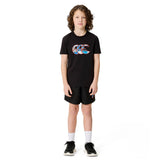 A child with curly hair stands facing forward against a white background, dressed in the Canterbury Youth Uglies Logo T-shirt in black, which showcases a vibrant Canterbury logo on the front. The t-shirt is paired with black shorts, white socks, and black sneakers. Made from comfortable cotton fabric, it offers all-day ease for active kids.