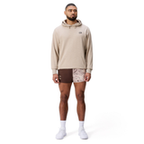 A man faces forward against a white background, wearing the Canterbury FORCE HOODIE in beige, patterned athletic shorts, white socks, and white sneakers.
