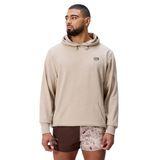 A man models the Canterbury FORCE HOODIE in beige, paired with brown and pink patterned shorts, standing slightly turned left against a plain background.