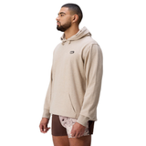 A man stands facing left against a plain white background, wearing a beige Canterbury FORCE HOODIE and brown athletic shorts.