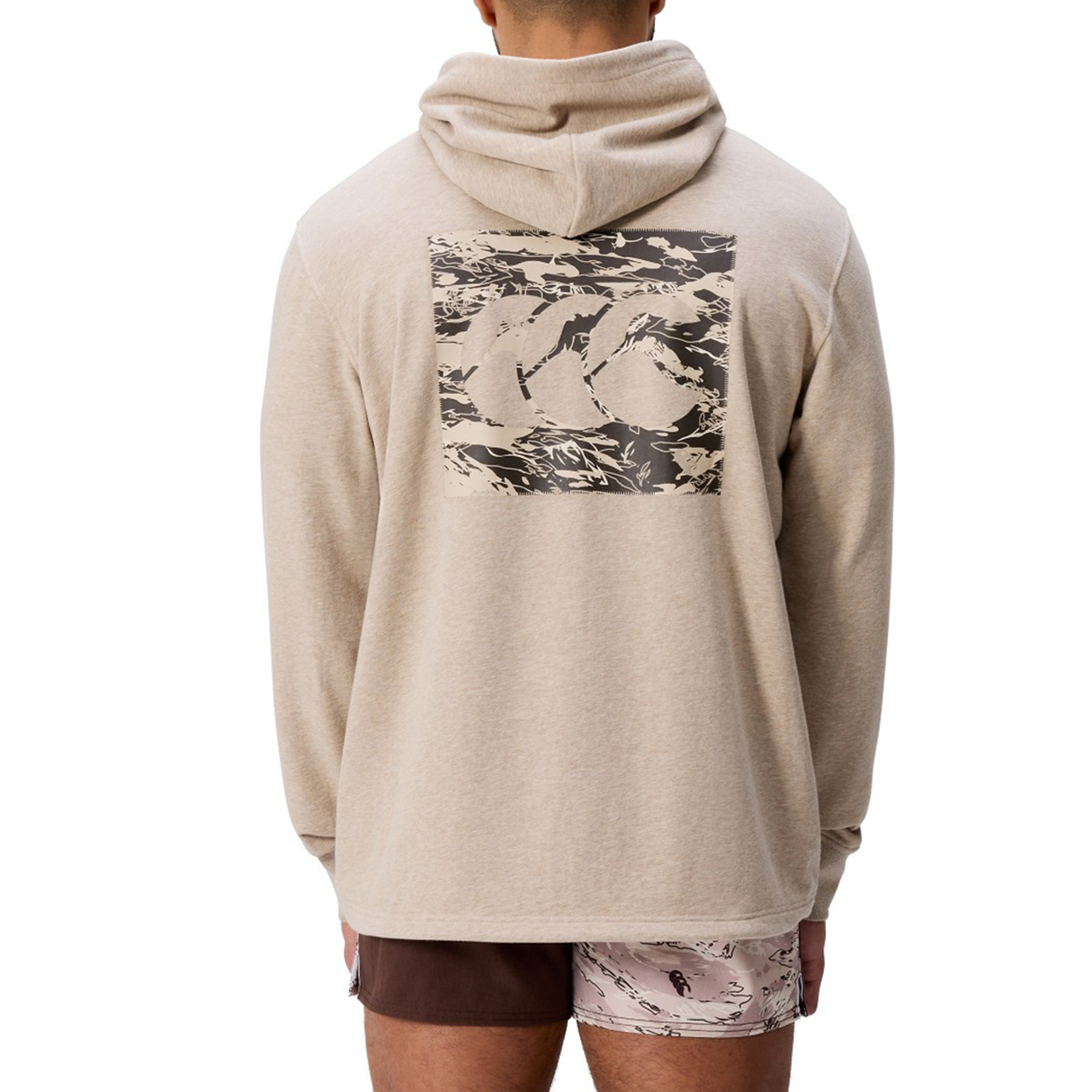 Someone is seen from behind wearing the Canterbury FORCE HOODIE in beige, featuring a large abstract graphic on the back, paired with two-tone shorts.