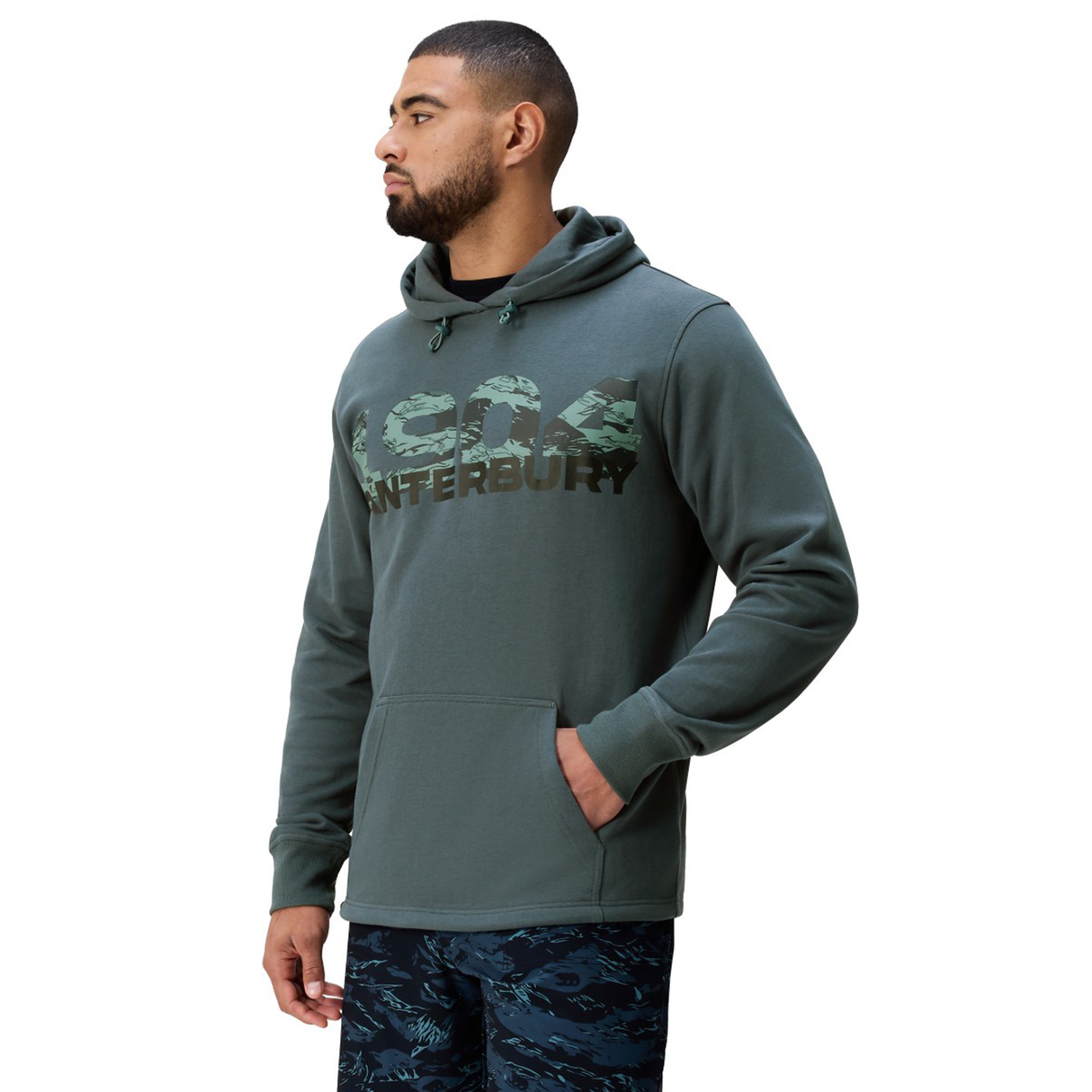 A man wearing a green Canterbury FORCE HOODIE and green patterned shorts stands facing left with one hand in his pocket.