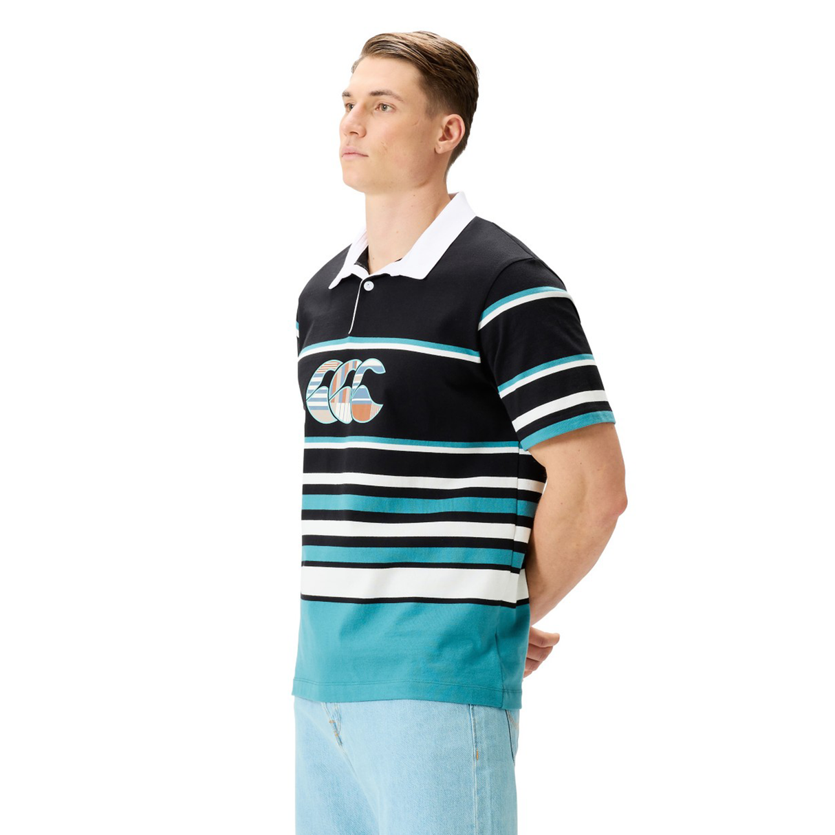 A man in a Canterbury Uglies Short Sleeve Classic Rugby Jersey featuring bold stripes and a large chest logo stands with his hands behind his back, looking off to the side.