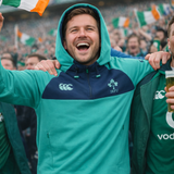A man in a Canterbury Ireland 26/27 Summer Tour Training Hoodie smiles with arms raised as a cheering crowd with Irish flags surrounds him at a sports event.