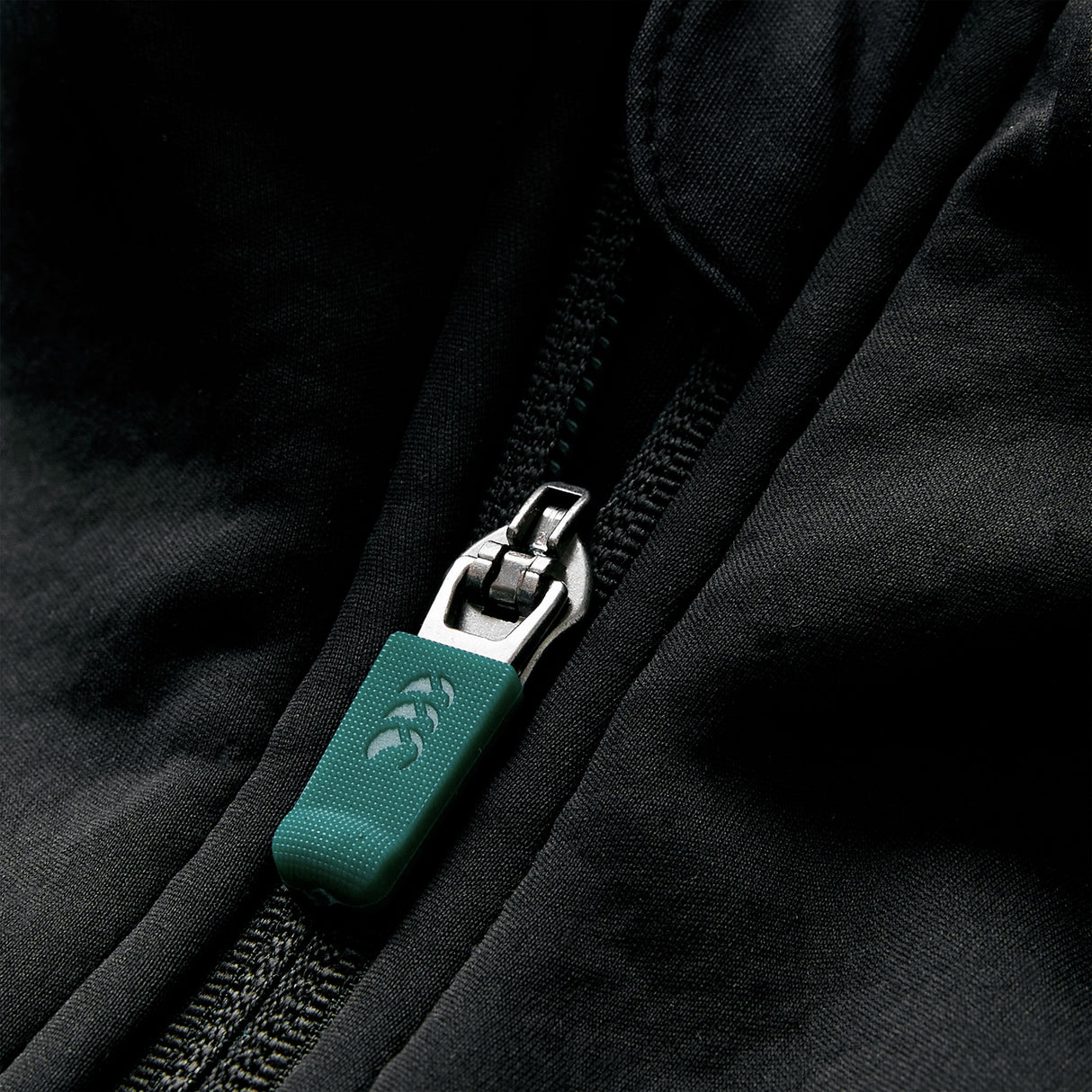 Close-up of a green and silver zipper on black fabric, with the zipper partially closed and the fabric slightly wrinkled. This Ireland 26 1/4 Zip Training Top by Canterbury showcases innovative Thermoreg technology for enhanced comfort during rugby training.