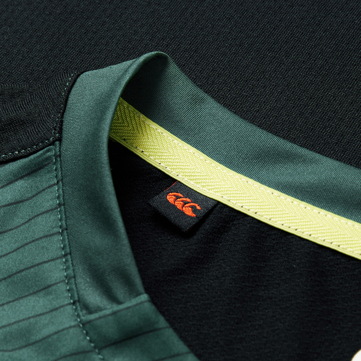 Close-up of the Ireland 26 Training Tank by Canterbury AW26 sports jersey collar in green, featuring a yellow stripe and a black tag with an orange Canterbury logo.