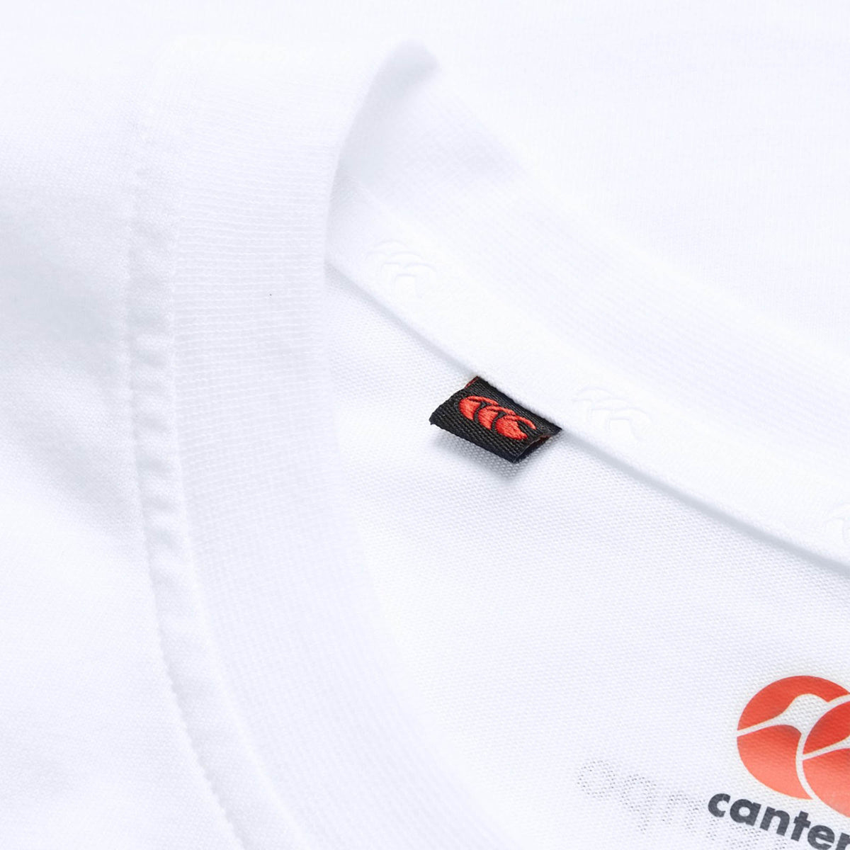 Close-up of the neckline on the Canterbury Tempo+ GFX Training Tank in white, featuring a small black tag with a red Canterbury logo. A partial orange graphic is visible on the fabric, highlighting the meticulous design that makes Canterbury training wear ideal for rugby fans seeking both style and performance.