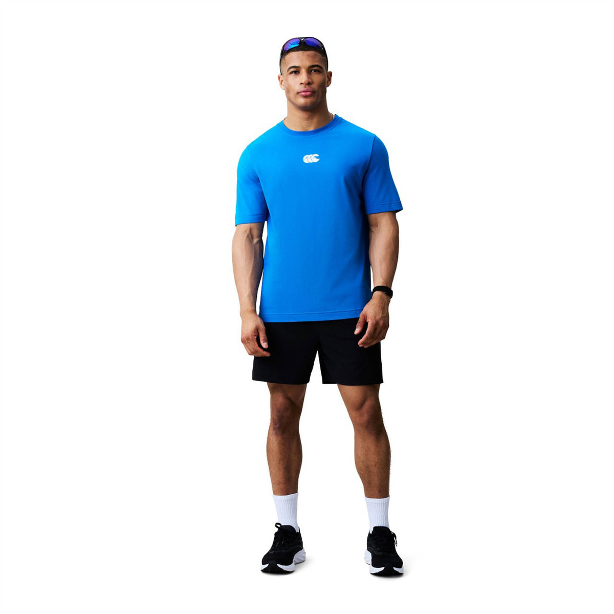 A man stands against a white background wearing a blue Canterbury Tempo+ GFX Training T-Shirt by Canterbury, paired with black shorts, black shoes, white socks, a watch on his wrist, and sunglasses resting on his head.