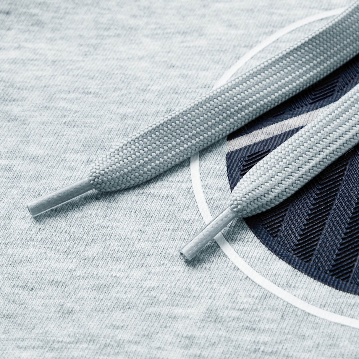 Close-up of two light gray shoelaces lying on heather gray fabric with a partial dark blue circular graphic, as featured on the Women Rakaia 2.0 Hoodie by Canterbury—a classic women’s rugby hoodie offering both comfort and style from Canterbury.