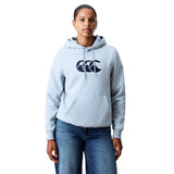 A person stands facing forward, wearing a light gray Women Rakaia 2.0 Hoodie by Canterbury featuring a dark logo, paired with blue jeans, against a plain white background.