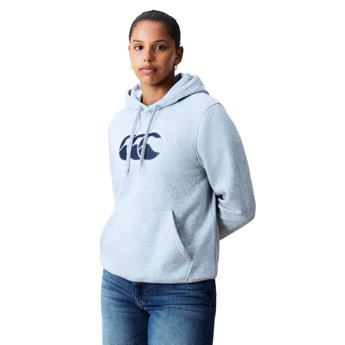 A person wearing a light gray Canterbury Women Rakaia 2.0 Hoodie with a front pocket and logo, paired with blue jeans, stands facing forward against a white background.