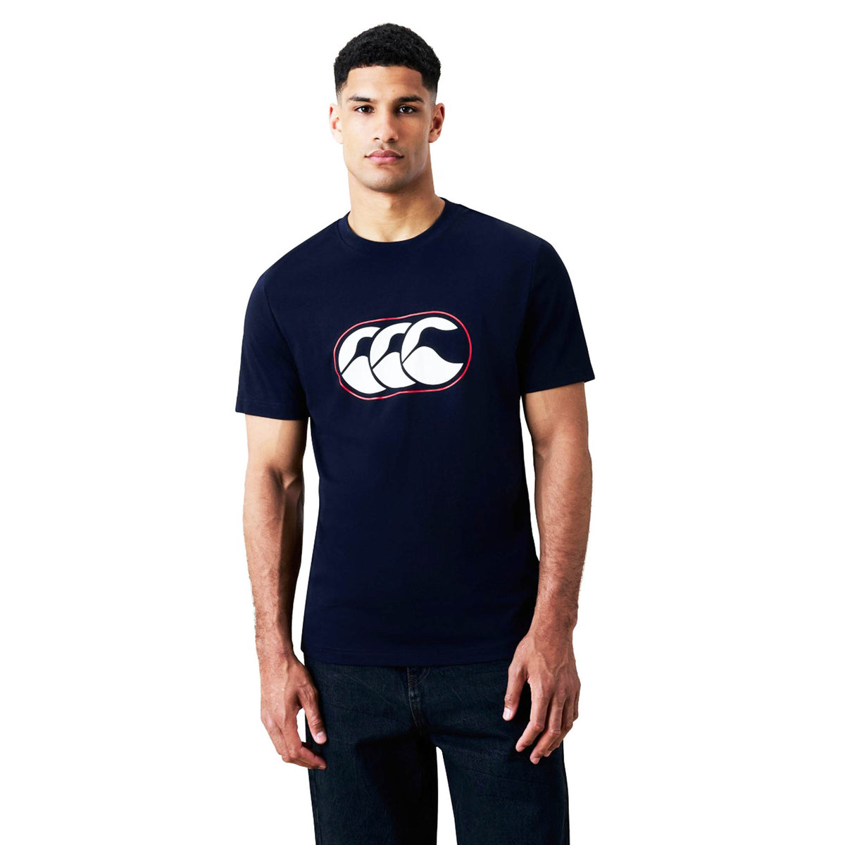 A man stands against a plain white background wearing a navy Canterbury Rakaia 2.0 T-shirt, featuring a white and red graphic logo on the chest and paired with dark jeans, embodying signature Canterbury New Zealand rugby apparel style.