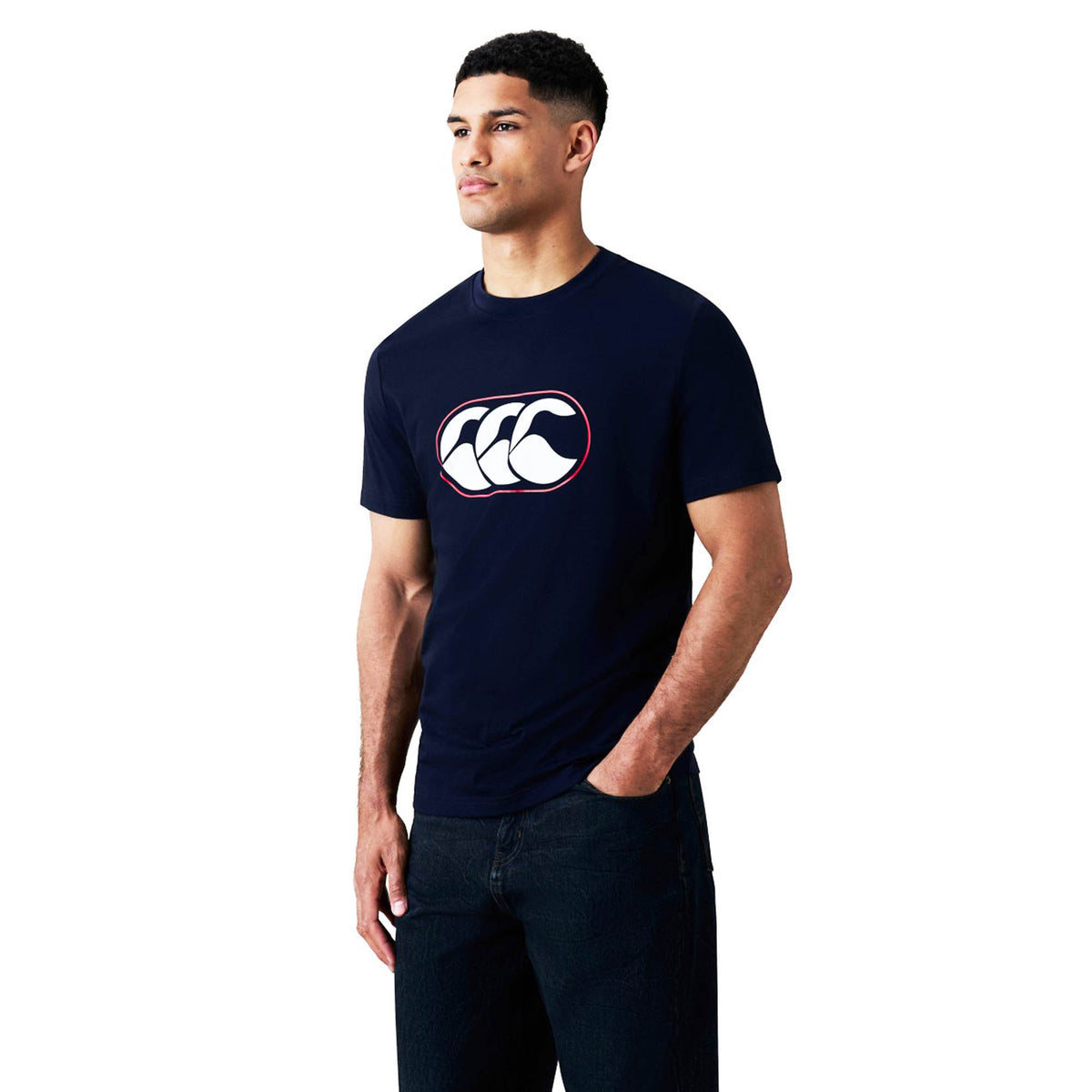 A man stands with one hand in his pocket against a white background, dressed in the Canterbury Rakaia 2.0 T-shirt by Canterbury, featuring a navy blue design with a white and red logo on the chest—an excellent pick for fans of New Zealand rugby apparel.