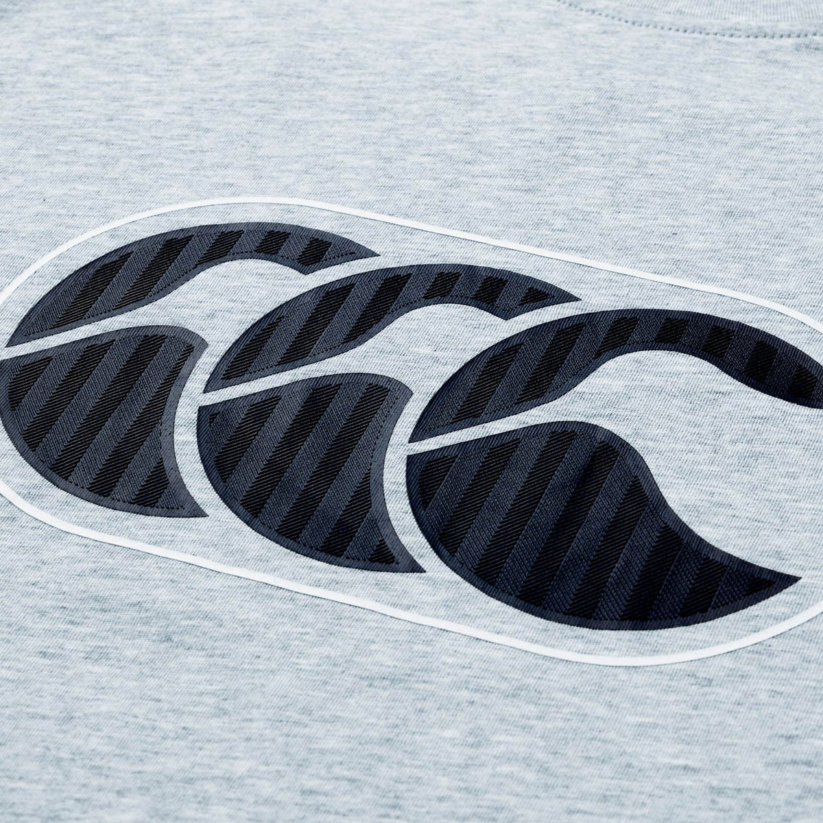 Close-up of a gray fabric from the Canterbury Rakaia 2.0 T-shirt, featuring a prominent dark blue Canterbury logo with three overlapping stylized C shapes—a hallmark of authentic New Zealand rugby apparel.
