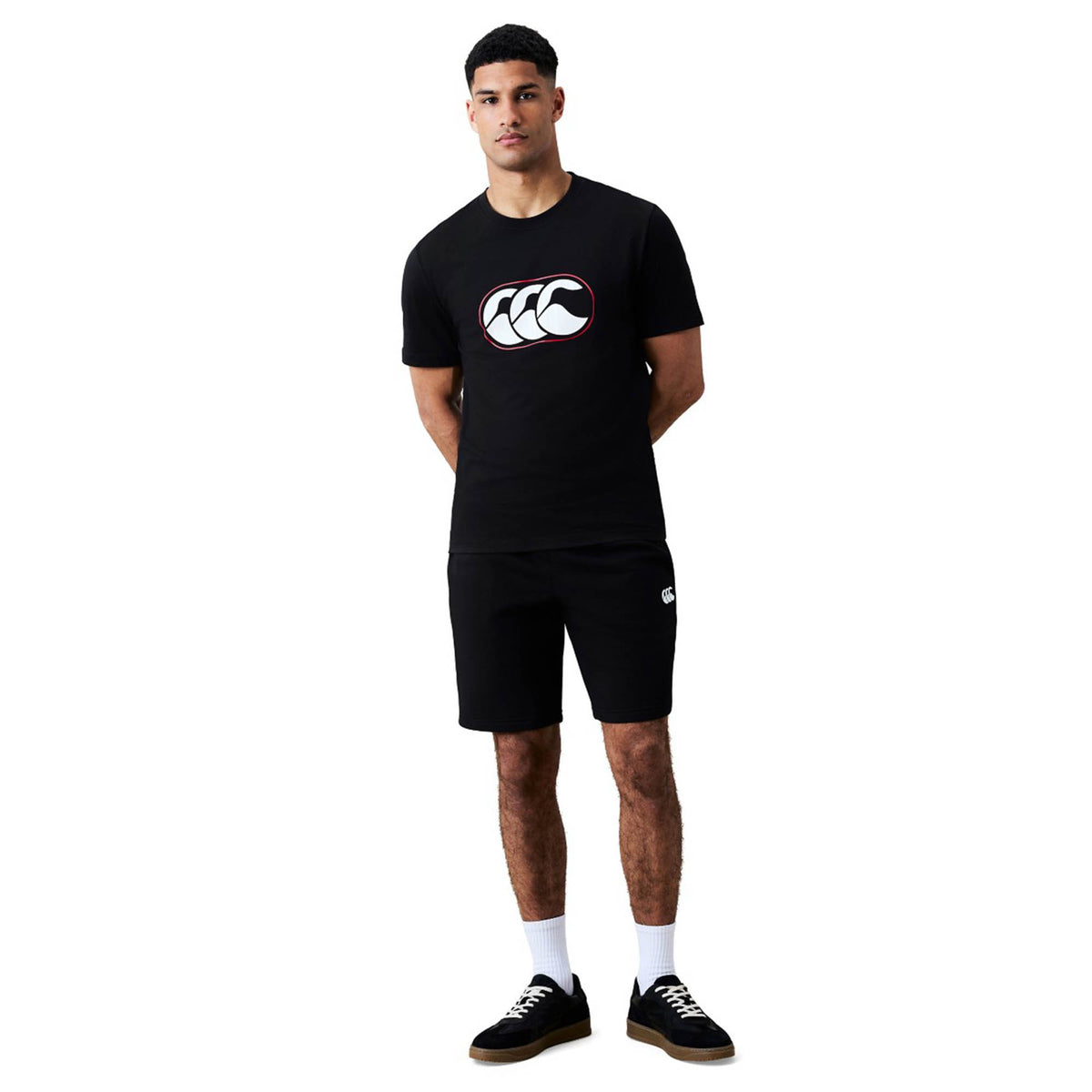A man stands against a white background wearing a black Canterbury Rakaia 2.0 T-shirt featuring a white logo, paired with black shorts, white socks, and black sneakers—showcasing signature Canterbury New Zealand rugby apparel.