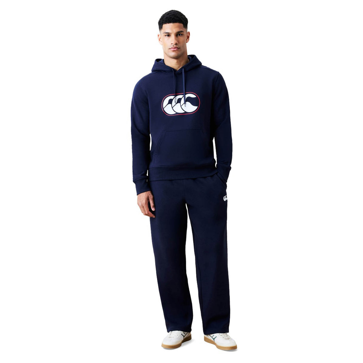 A man is wearing the Canterbury Rakaia 2.0 Hoodie by Canterbury in navy blue, featuring the brand logo, paired with matching navy blue sweatpants and white sneakers, standing against a plain white background.