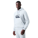 A man stands facing forward, wearing a matching set of light grey Canterbury Rakaia 2.0 Hoodie and sweatpants. The Canterbury rugby hoodie is detailed with a black logo graphic on the chest.