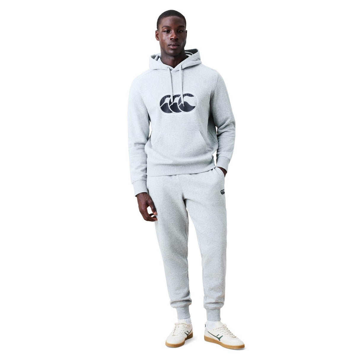 A man stands wearing a light gray Canterbury Rakaia 2.0 Hoodie and matching sweatpants featuring the Canterbury logo on the chest, paired with white sneakers, against a plain white background.