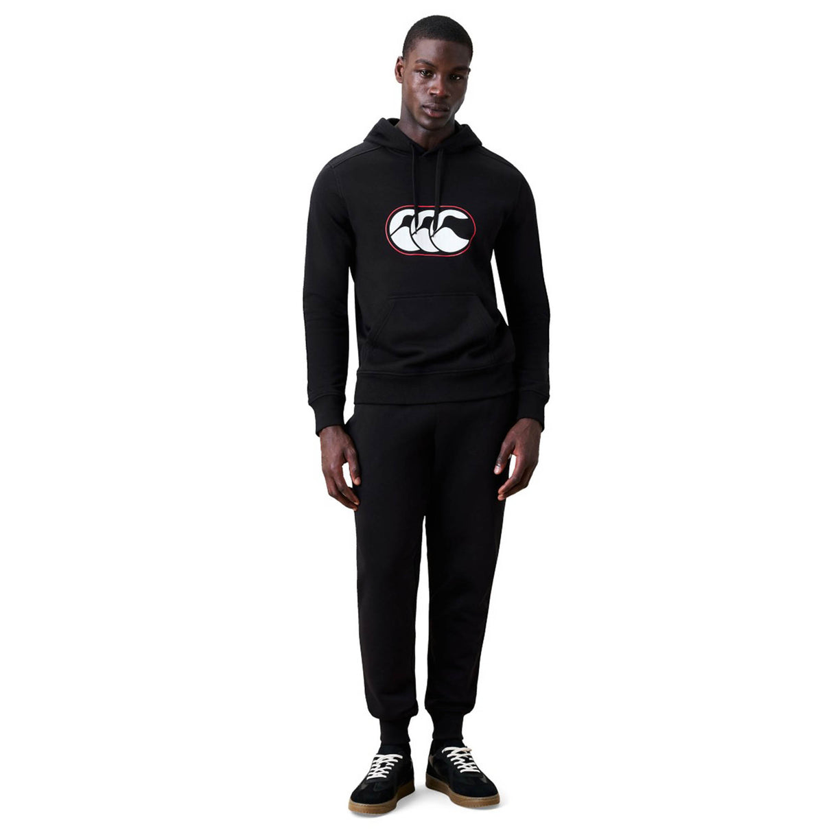 A man stands against a white background wearing the Canterbury Rakaia 2.0 Hoodie by Canterbury, featuring a black design with white and red logo, paired with black sweatpants and black sneakers.