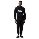 A man stands against a white background wearing the Canterbury Rakaia 2.0 Hoodie by Canterbury, featuring a black design with white and red logo, paired with black sweatpants and black sneakers.