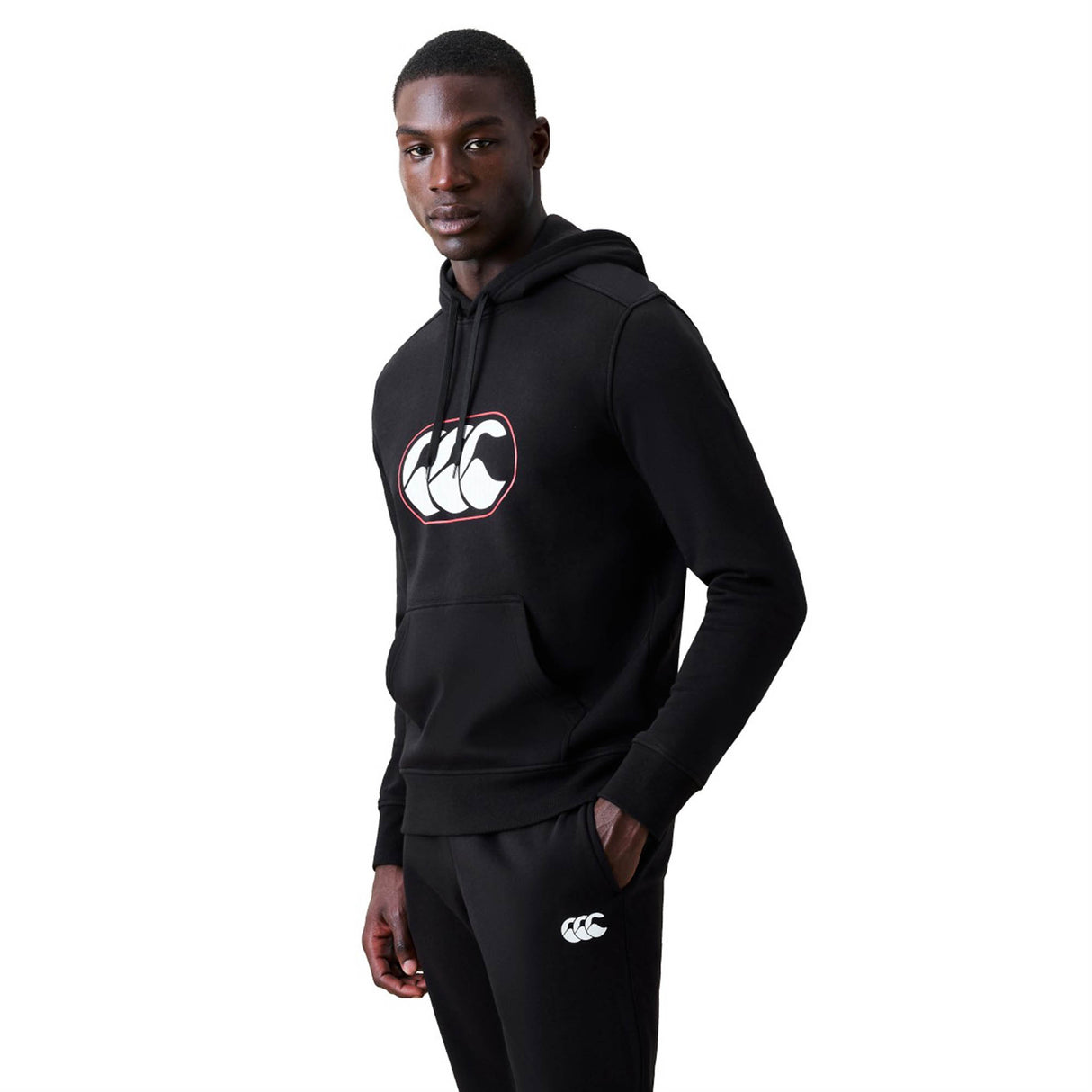 A person wearing a black Canterbury Rakaia 2.0 Hoodie and matching black pants with white Canterbury logos stands facing slightly to the side against a plain white background.