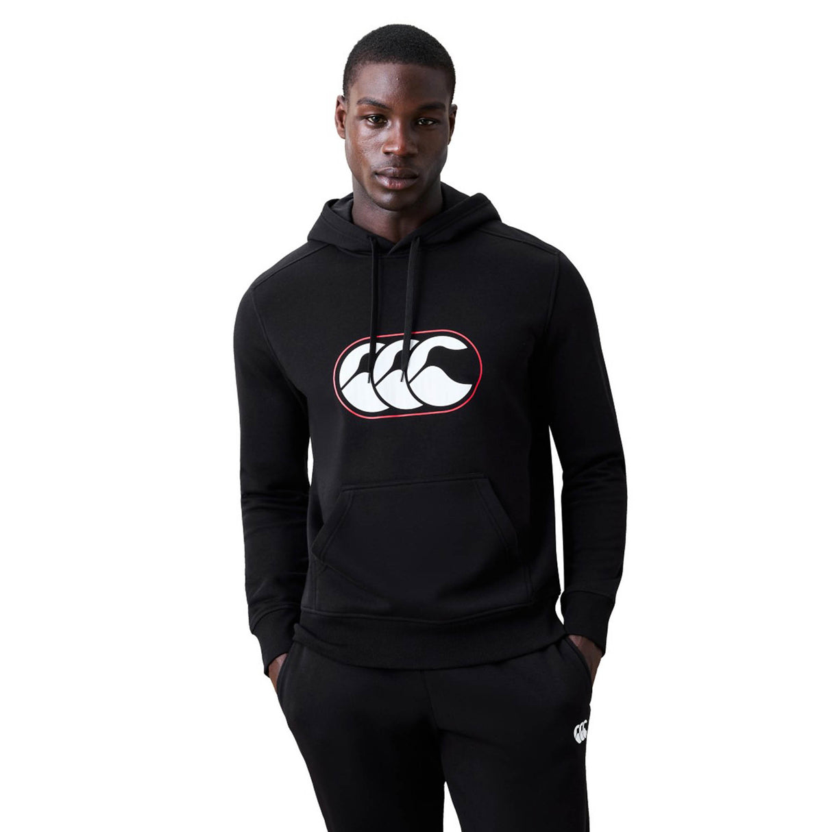 A man wearing the Canterbury Rakaia 2.0 Hoodie by Canterbury, featuring a black design with a white and red logo on the front, stands with his hands in his pockets against a plain background.