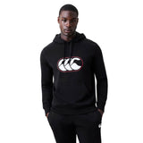 A man wearing the Canterbury Rakaia 2.0 Hoodie by Canterbury, featuring a black design with a white and red logo on the front, stands with his hands in his pockets against a plain background.