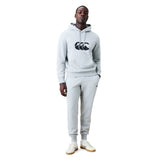 A person stands against a plain white background wearing a light gray hoodie with a black logo, paired with matching Canterbury Rakaia 2.0 Fleece Pants and white sneakers.