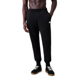 A person wearing the black Rakaia 2.0 Fleece Pant by Canterbury with hands in pockets, paired with black sneakers featuring white laces, stands against a white background.