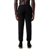 A person wearing black Rakaia 2.0 Fleece Pants by Canterbury and black sneakers stands facing away against a white background.