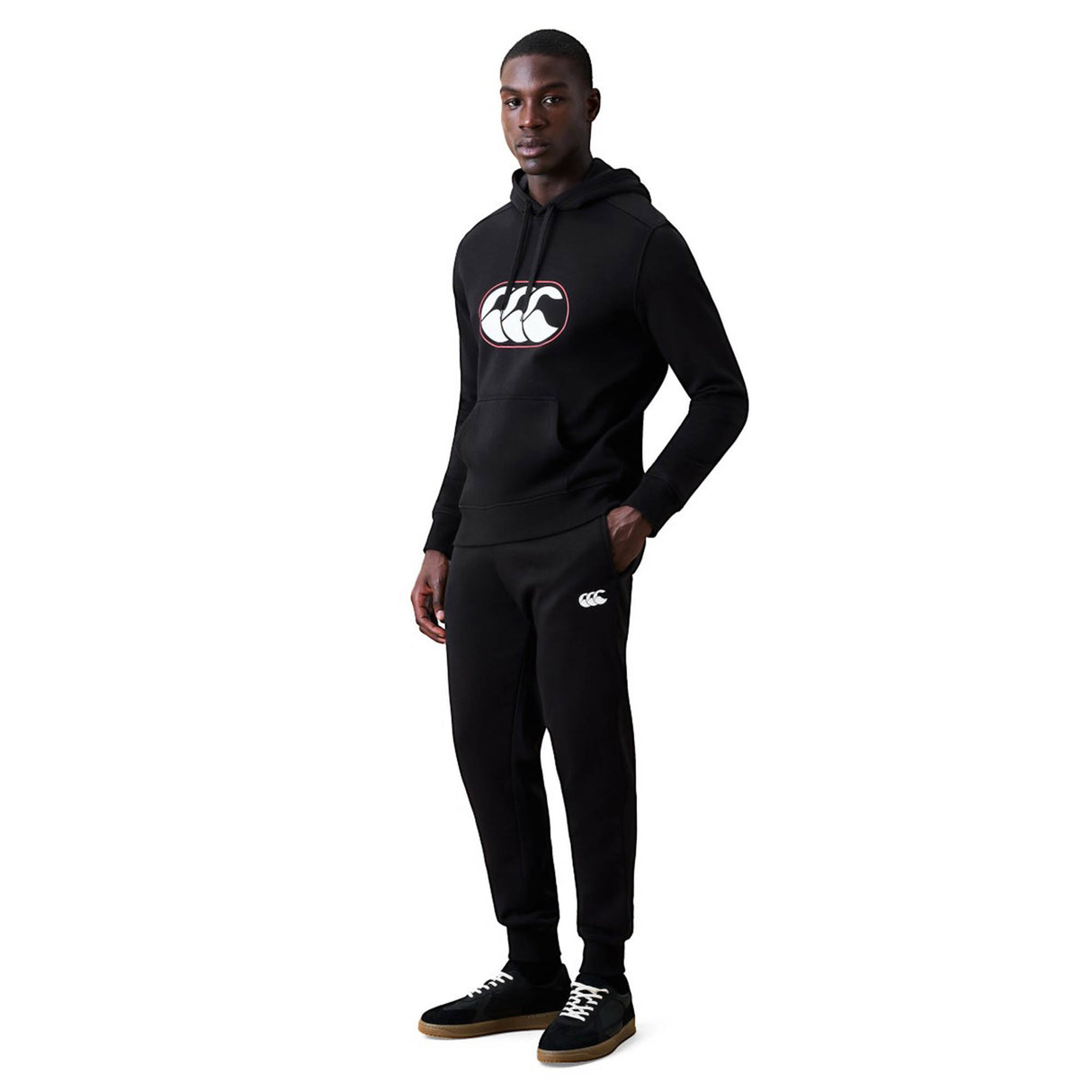 A person wearing a black hooded sweatshirt and matching Canterbury Rakaia 2.0 Fleece Pant featuring white logos stands with one hand in their pocket against a white background.