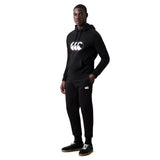A person wearing a black hooded sweatshirt and matching Canterbury Rakaia 2.0 Fleece Pant featuring white logos stands with one hand in their pocket against a white background.