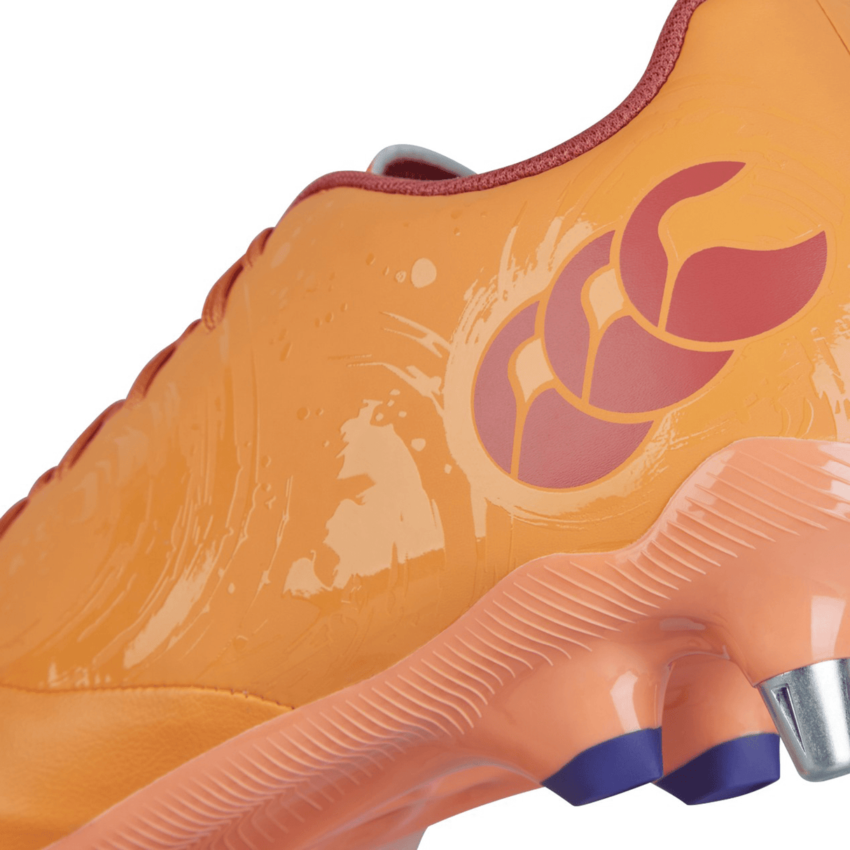 Close-up of an orange Canterbury Phoenix Genesis Pro SG sports cleat in the Orange Crush/Amber Yellow/Mars Dust colorway, featuring a textured surface and a red three-leaf logo on the side. The shoe has a translucent orange sole with purple-tipped studs, offering ultimate traction on soft ground.