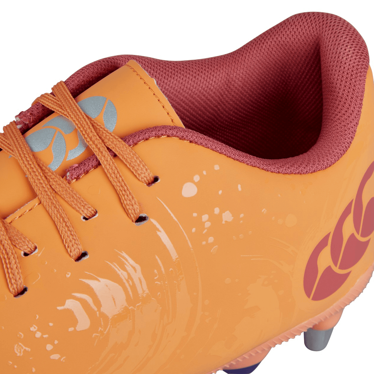 Close-up of the Canterbury Phoenix Genesis Pro SG in Orange Crush, Amber Yellow, and Mars Dust. This athletic shoe boasts a red lining and laces, a patterned design, and metal spikes on the sole for ultimate traction on soft ground terrain.